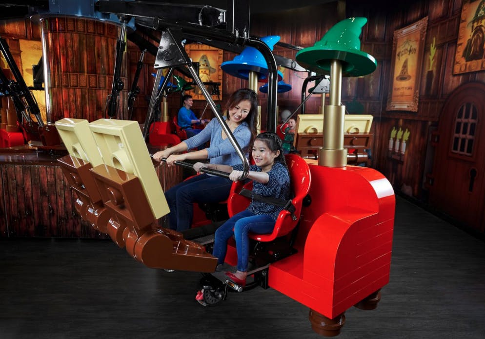 A mother and child enjoy a ride in a red and brown theme park vehicle that resembles a large LEGO brick.