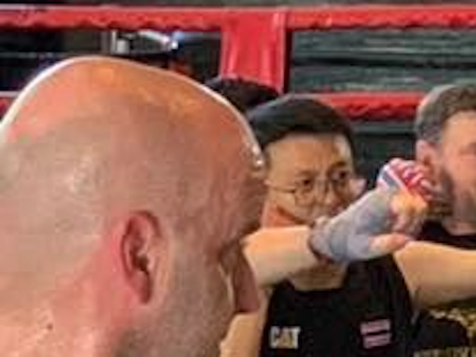 A group of people practicing martial arts in a gym, wearing matching black and gold uniforms, with a boxing ring in the background.