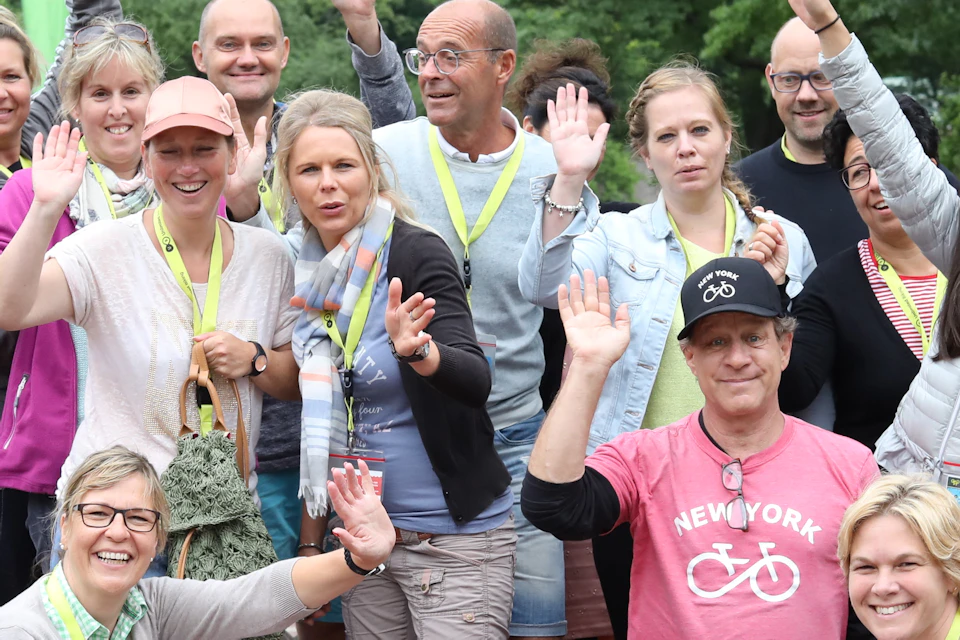 A group of smiling people posing and waving outdoors, some wearing badges and lanyards, with a backdrop of lush trees.