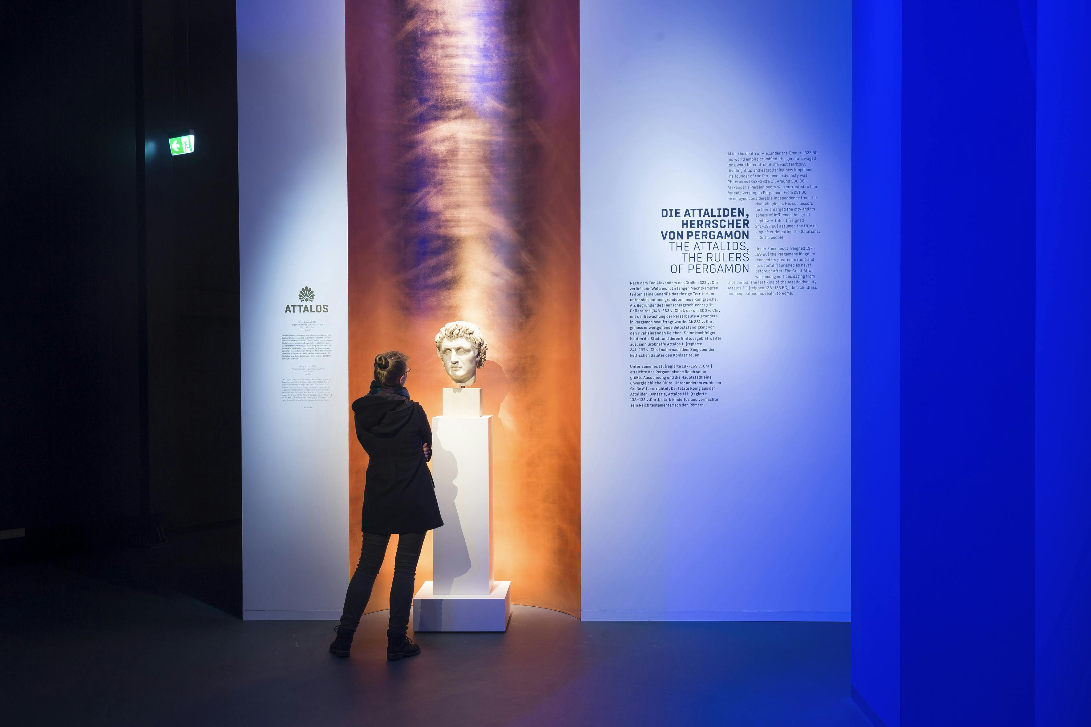 Osoba v černém kabátě stojí čelem k bílému podstavci s mramorovou bustou u zdi s textovými informacemi v tlumeně osvětlené místnosti.