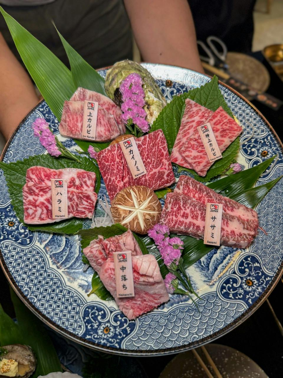 A decorative plate featuring an arrangement of marbled beef slices on leaves, garnished with flowers and a mushroom.