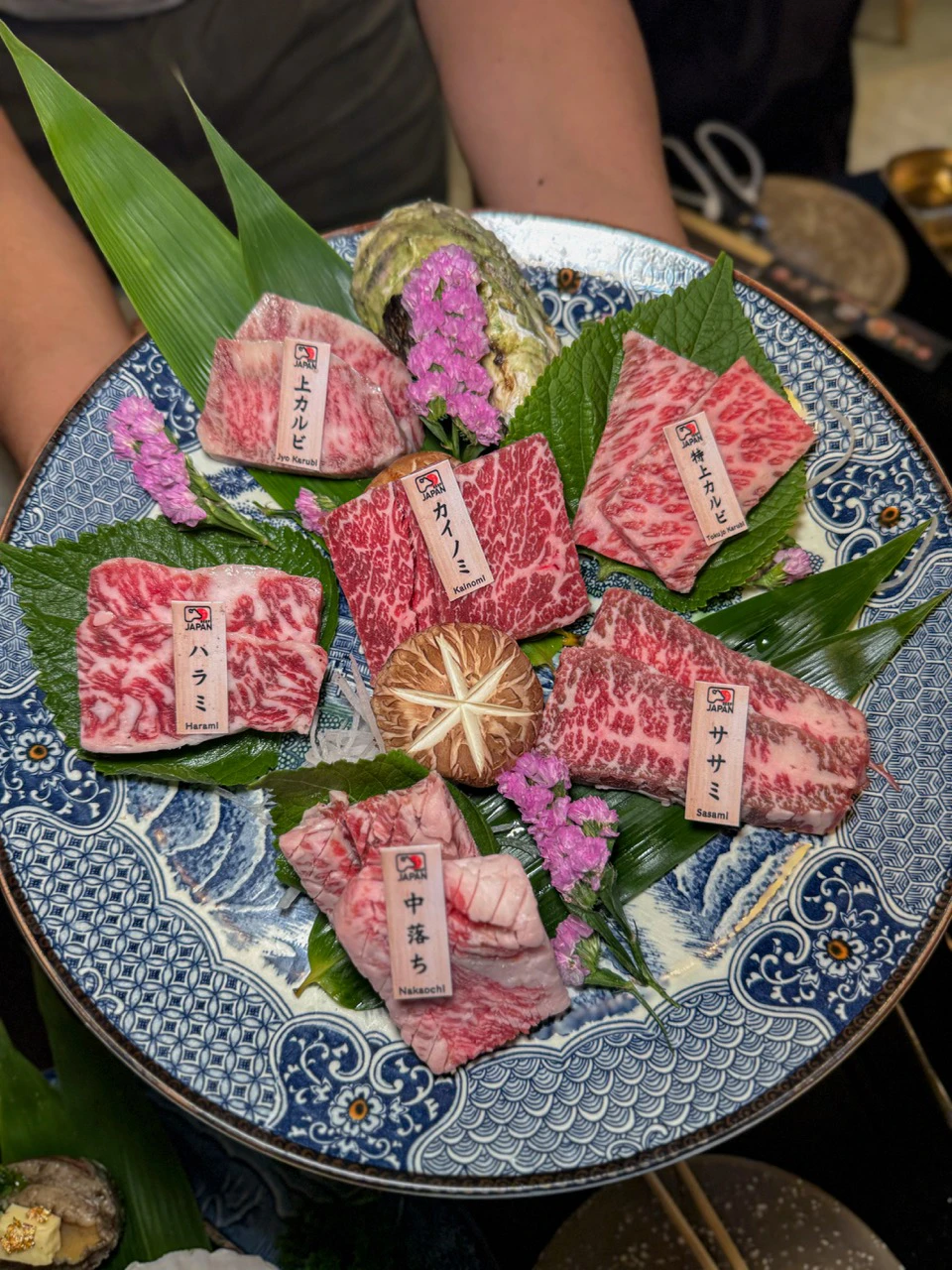A decorative plate featuring various cuts of marbled meat, garnished with green leaves and purple flowers.