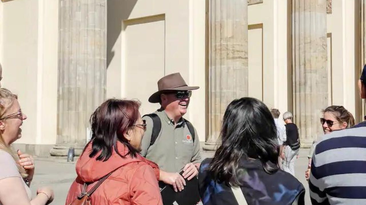 Turistický průvodce s kloboukem promlouvající ke skupině lidí před velkými kamennými sloupy za slunečného dne.