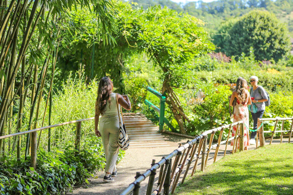 Two women walking on a sunlit path through a lush, green garden with wooden railings and an archway covered in foliage.