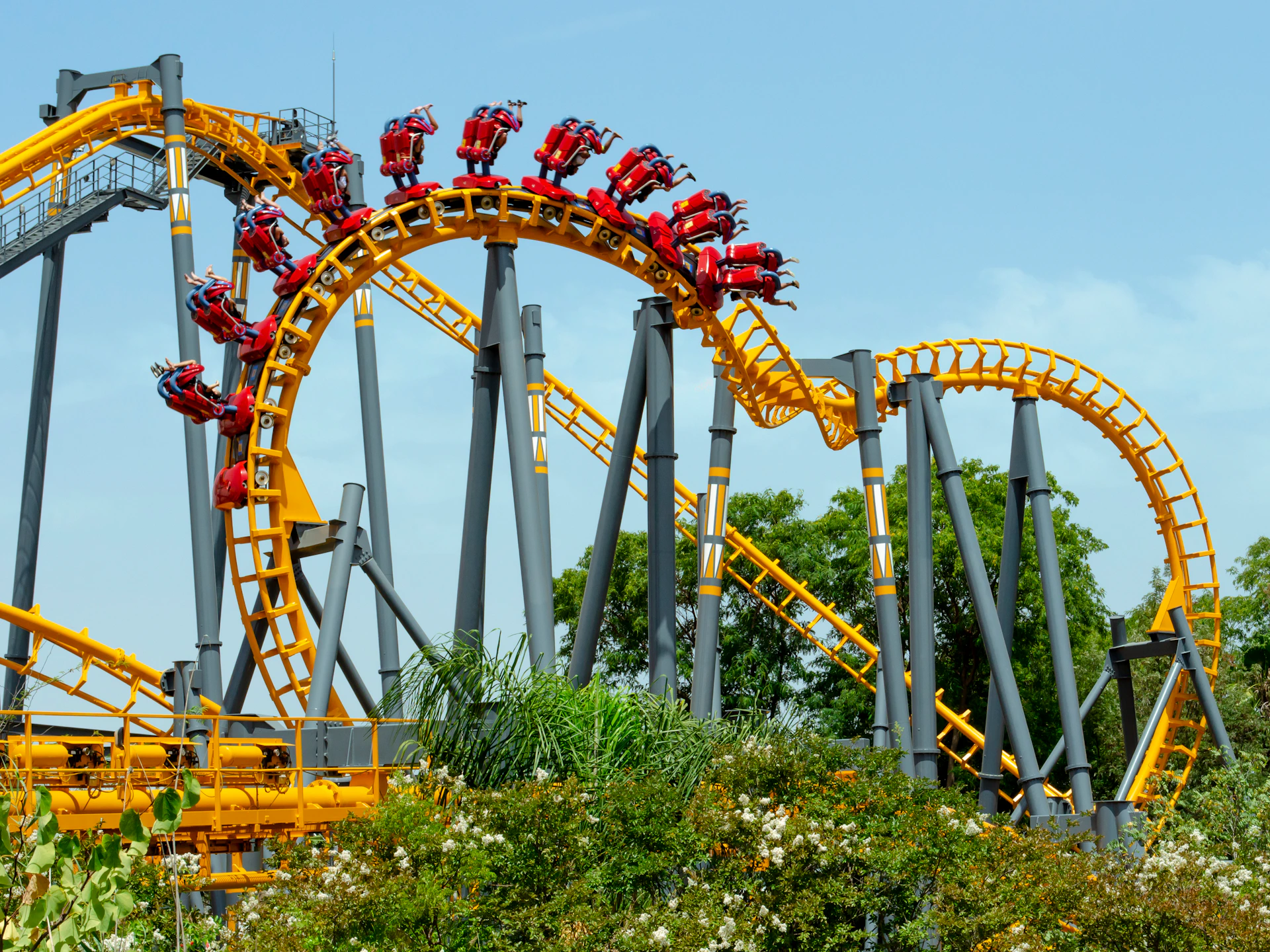 People riding a yellow and red rollercoaster with loops and turns, surrounded by greenery and a clear blue sky.