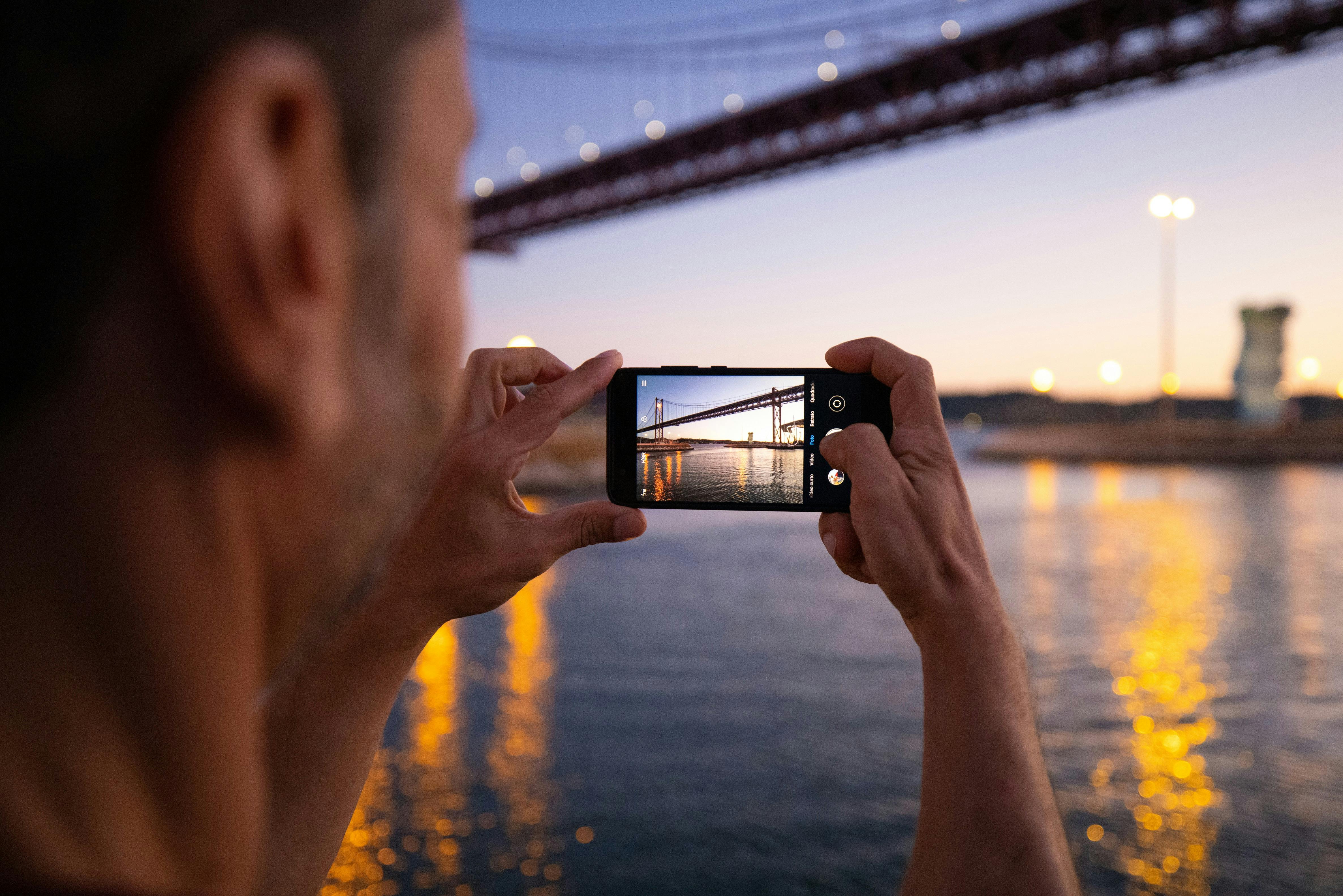 스마트폰을 들고 있는 사람이 해질녘 물 위에 있는 다리의 사진을 찍습니다.