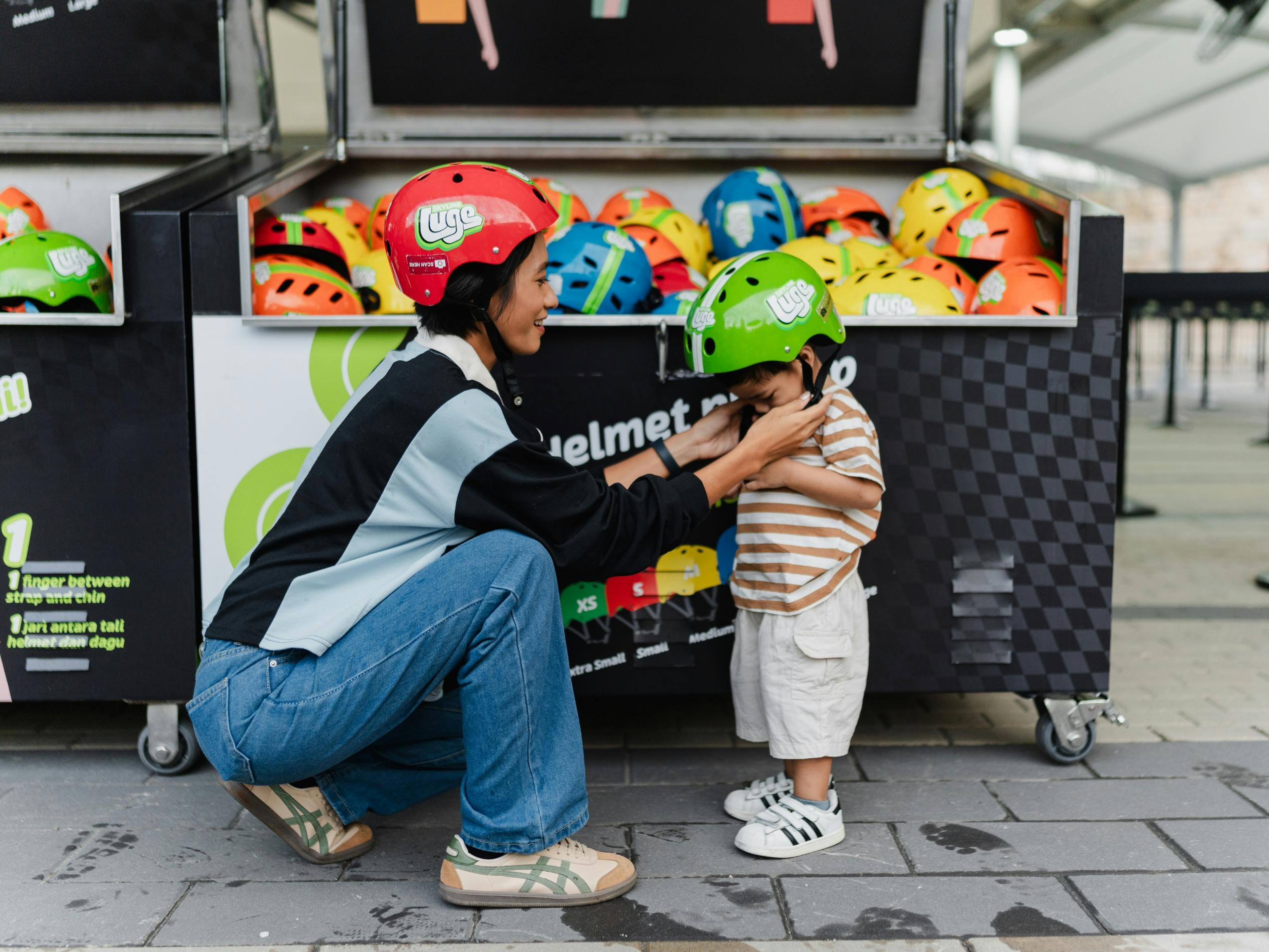 An adult helps a child adjust a green helmet in front of a display of colorful helmets. Both are smiling and interacting.