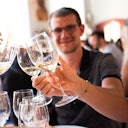A man wearing glasses clinks wine glasses with others at a social gathering, smiling and looking at the camera.