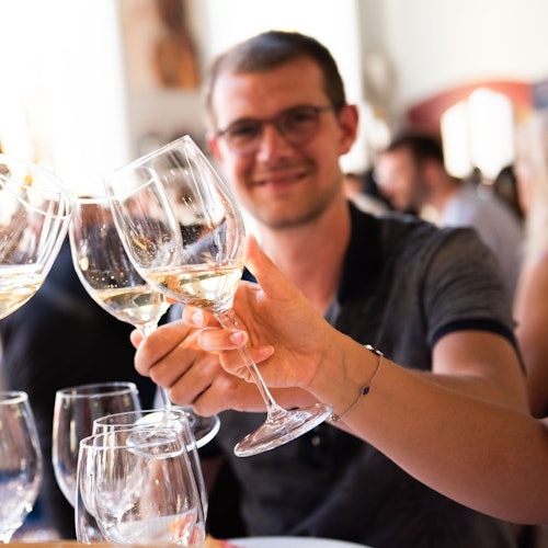 A man wearing glasses clinks wine glasses with others at a social gathering, smiling and looking at the camera.