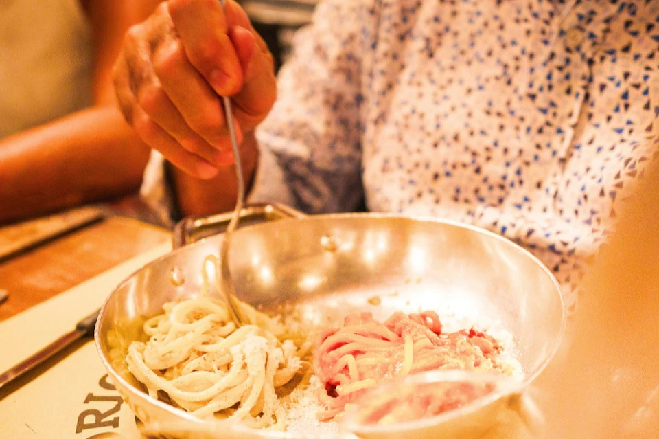 A person wearing a patterned shirt twirls pasta in a metallic pan with a fork.