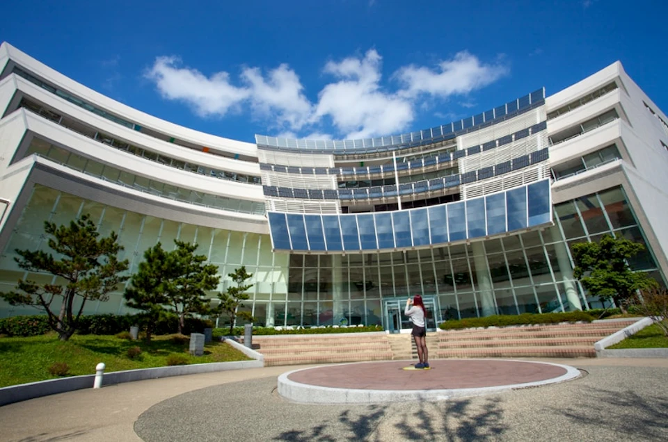 A person stands on a circular plaza in front of a modern, curved glass building under a blue sky with scattered clouds.