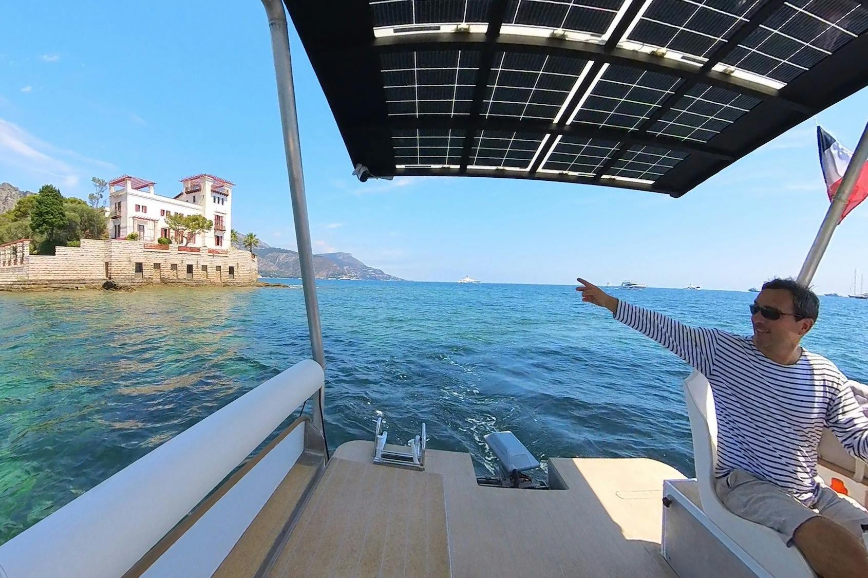 一名身穿条纹衬衫的男子坐在配备太阳能电池板的船上，指向海边的白色建筑。