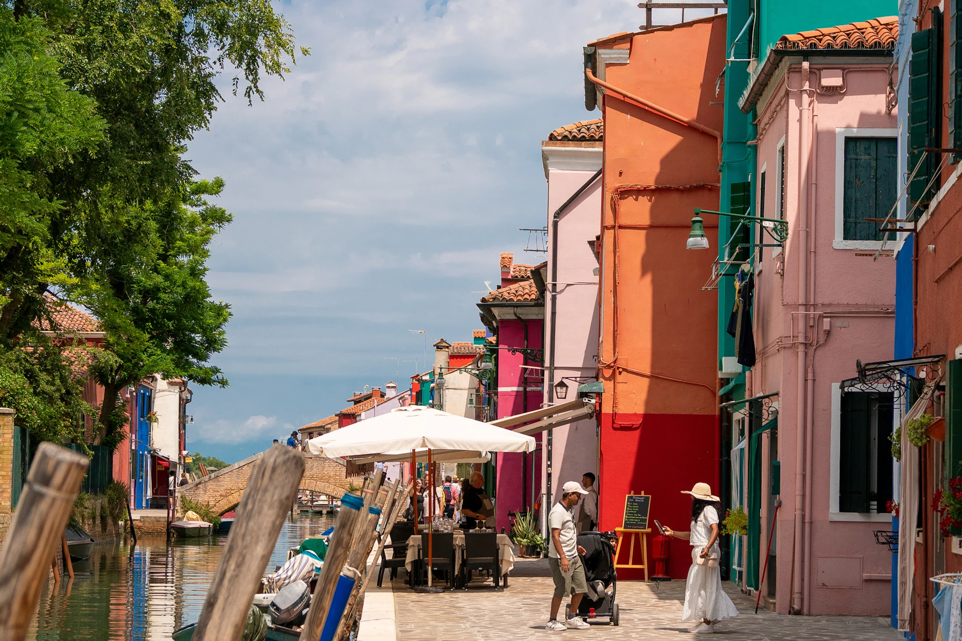People walking along a canal lined with colorful buildings, outdoor seating, and umbrellas under a partly cloudy sky.
