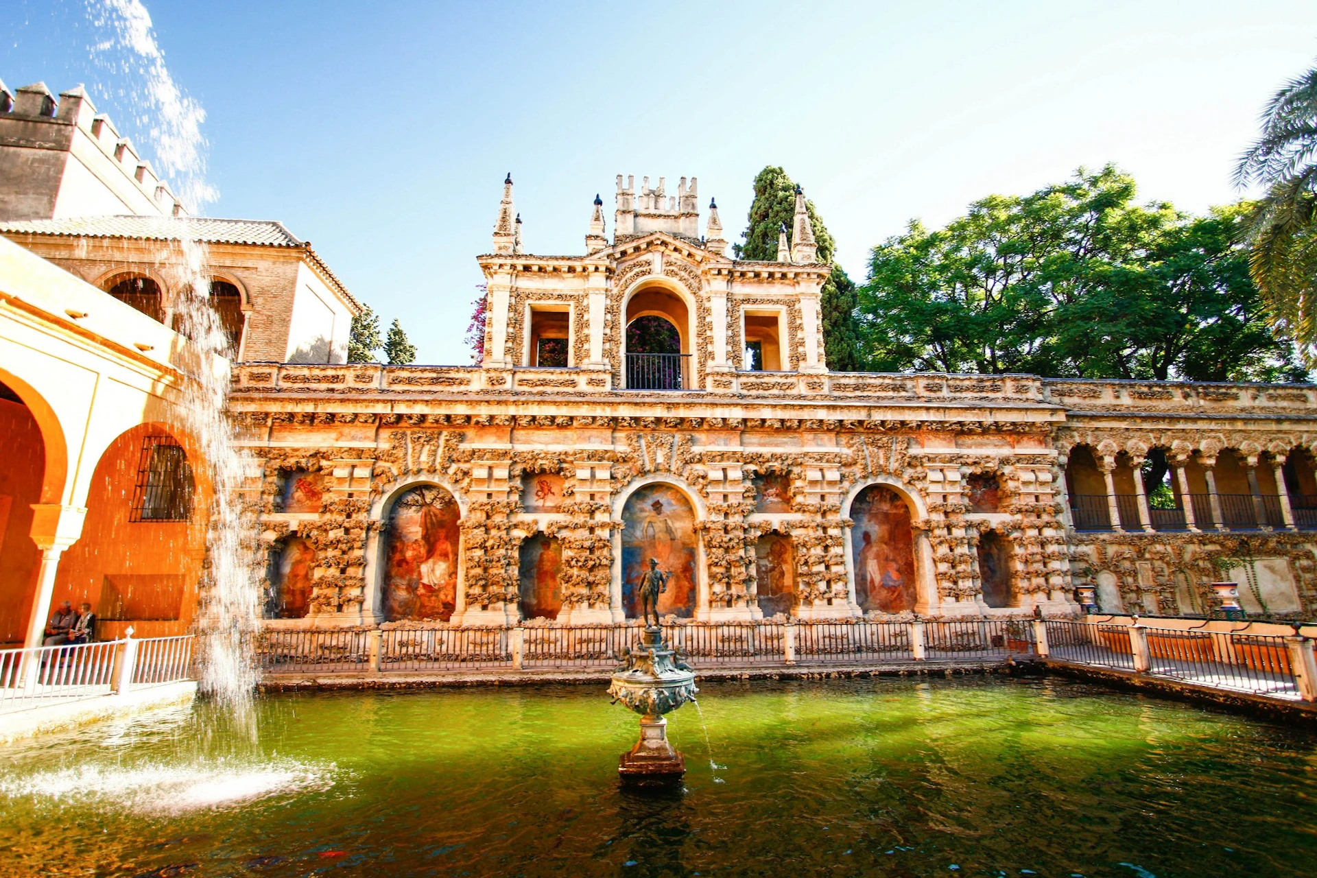 Ornate historic building with arches and statues reflected in a green pond, surrounded by trees under a clear sky.