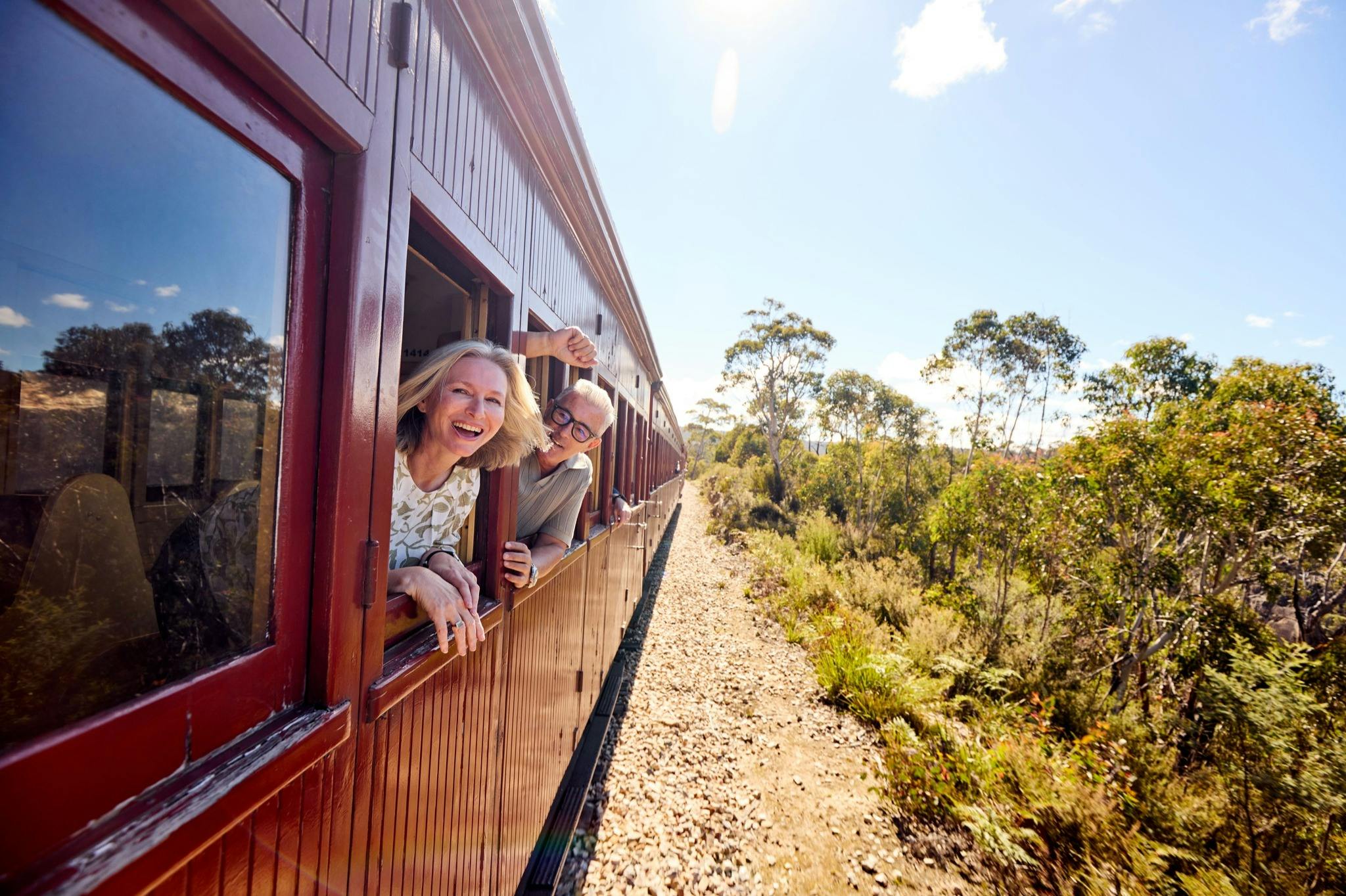 Two people smiling and leaning out of the windows of a red vintage train traveling through a sunny countryside.