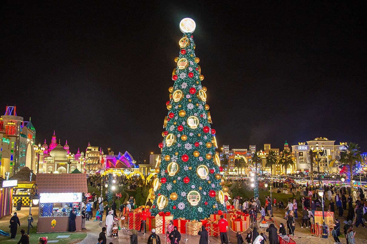 Velký, zdobený vánoční stromek s ornamenty a světly na rušném veřejném náměstí v noci. Kolem se shromažďují lidé, někteří ve slavnostním oděvu.