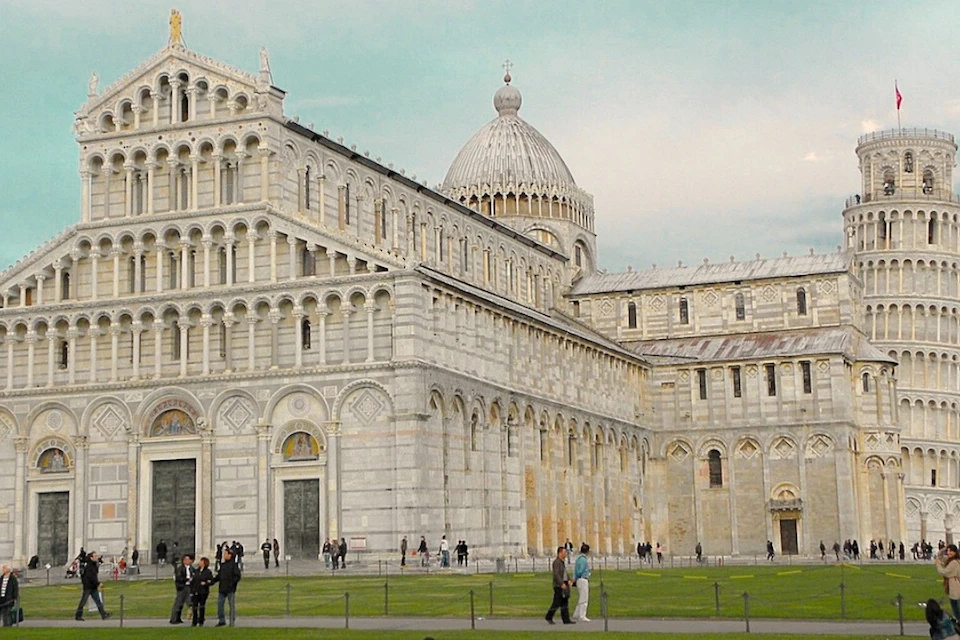A historic cathedral with intricate architecture, a domed roof, and people walking on the grass near the building.