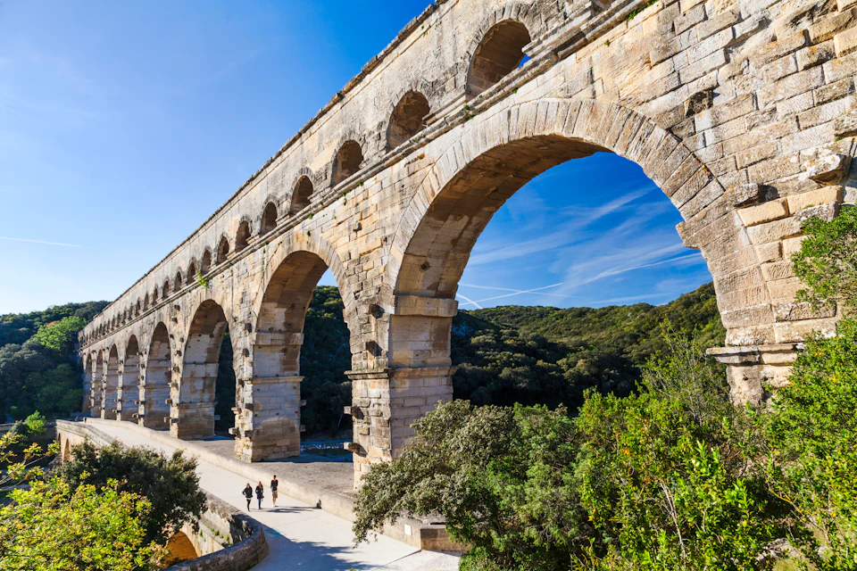 Ancient stone aqueduct with large arches set against a blue sky and green landscape. Three people are walking below.