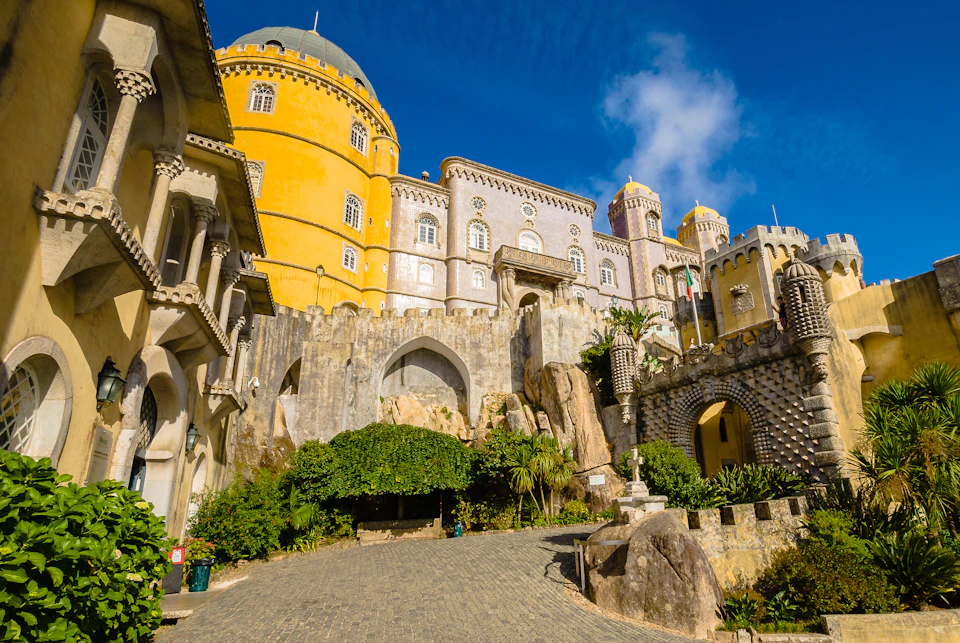 Vibrant yellow and grey castle with arched windows, domes, and lush greenery under a bright blue sky.