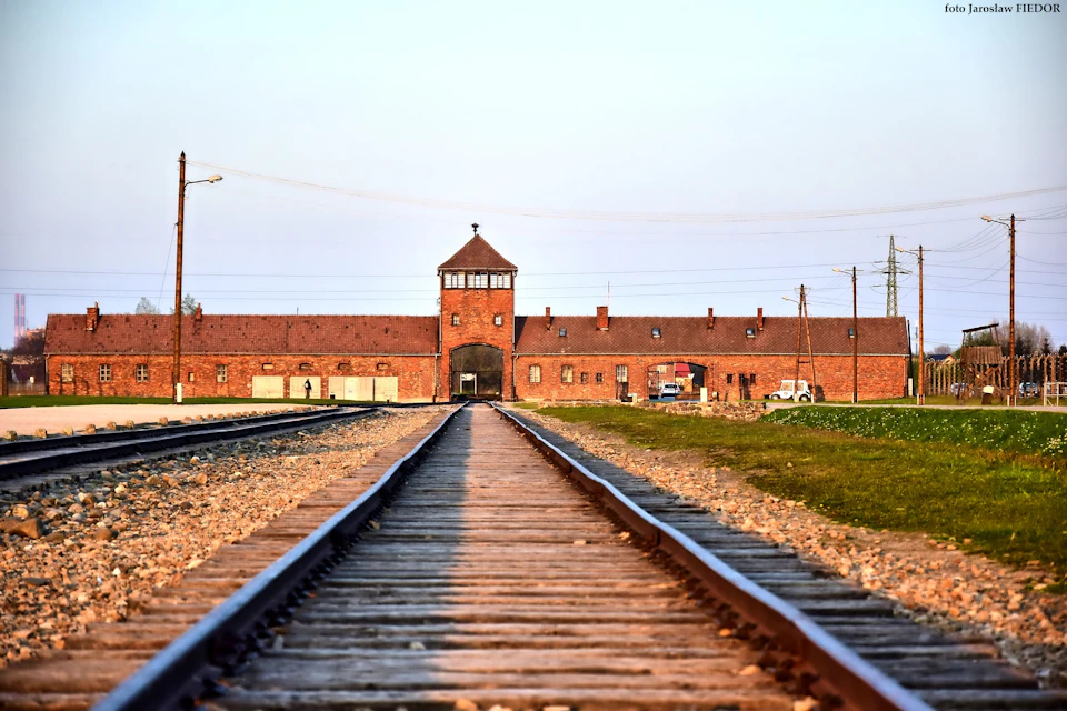 A railway track leading to a large brick building with an archway and a central tower under a clear sky.