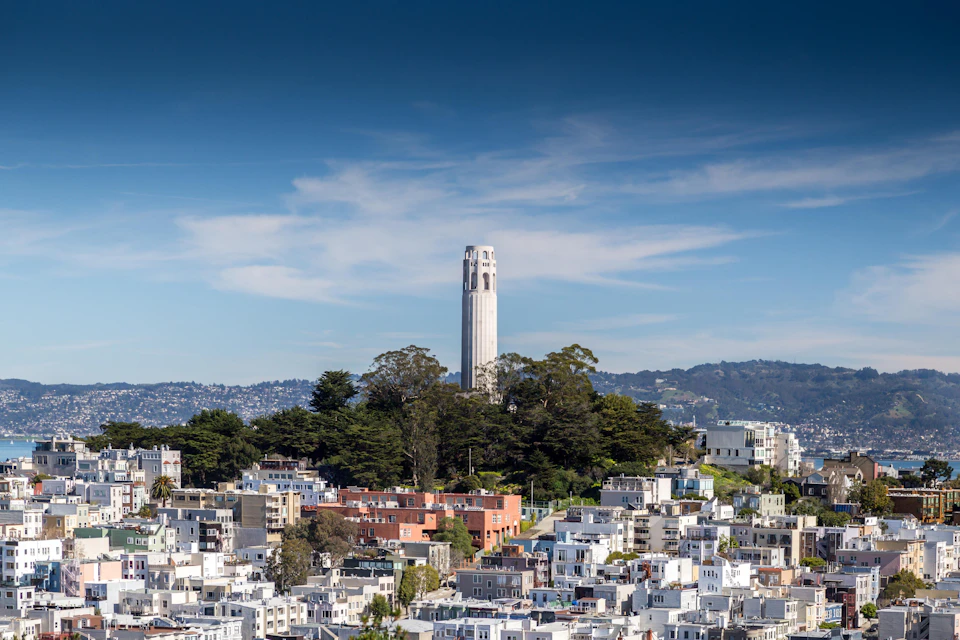 Tall tower surrounded by trees on a hill, with a cityscape of buildings below and mountains in the background under a clear sky.