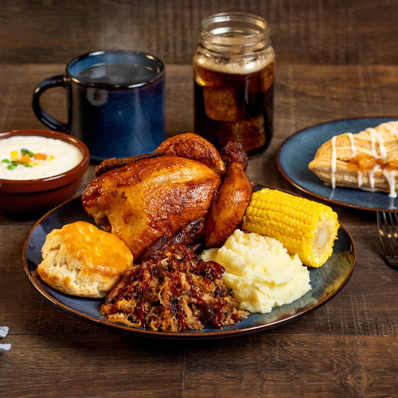 A meal featuring roasted chicken, corn, mashed potatoes, biscuit, and pulled pork on a plate, with sides and a drink.