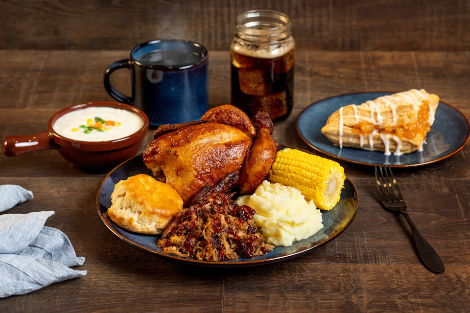 A meal featuring roasted chicken, corn, mashed potatoes, biscuit, and pulled pork on a plate, with sides and a drink.