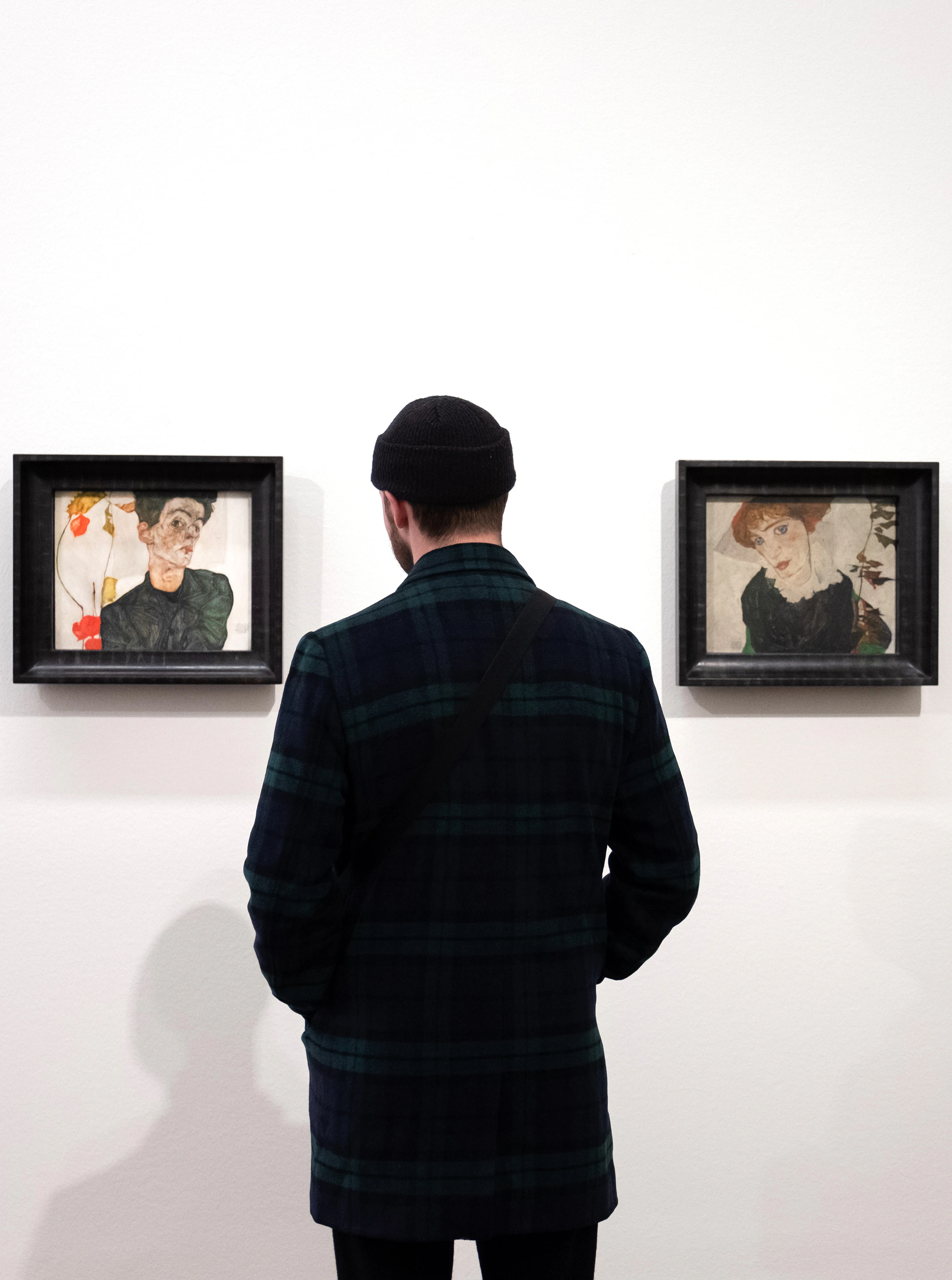 A person wearing a black beanie and dark plaid jacket stands in front of two framed artworks on a white wall.