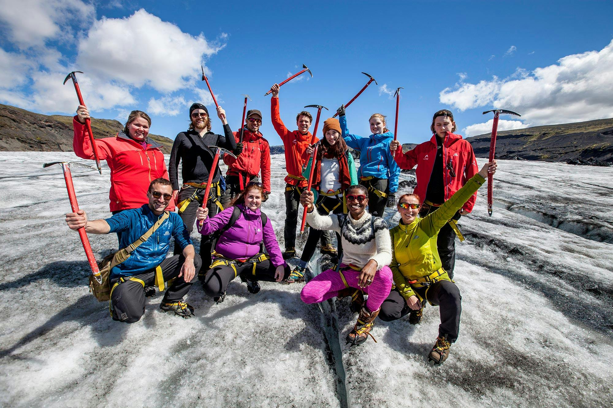 A group of smiling hikers in bright outdoor gear raises ice axes while standing on a glacier under a clear blue sky.