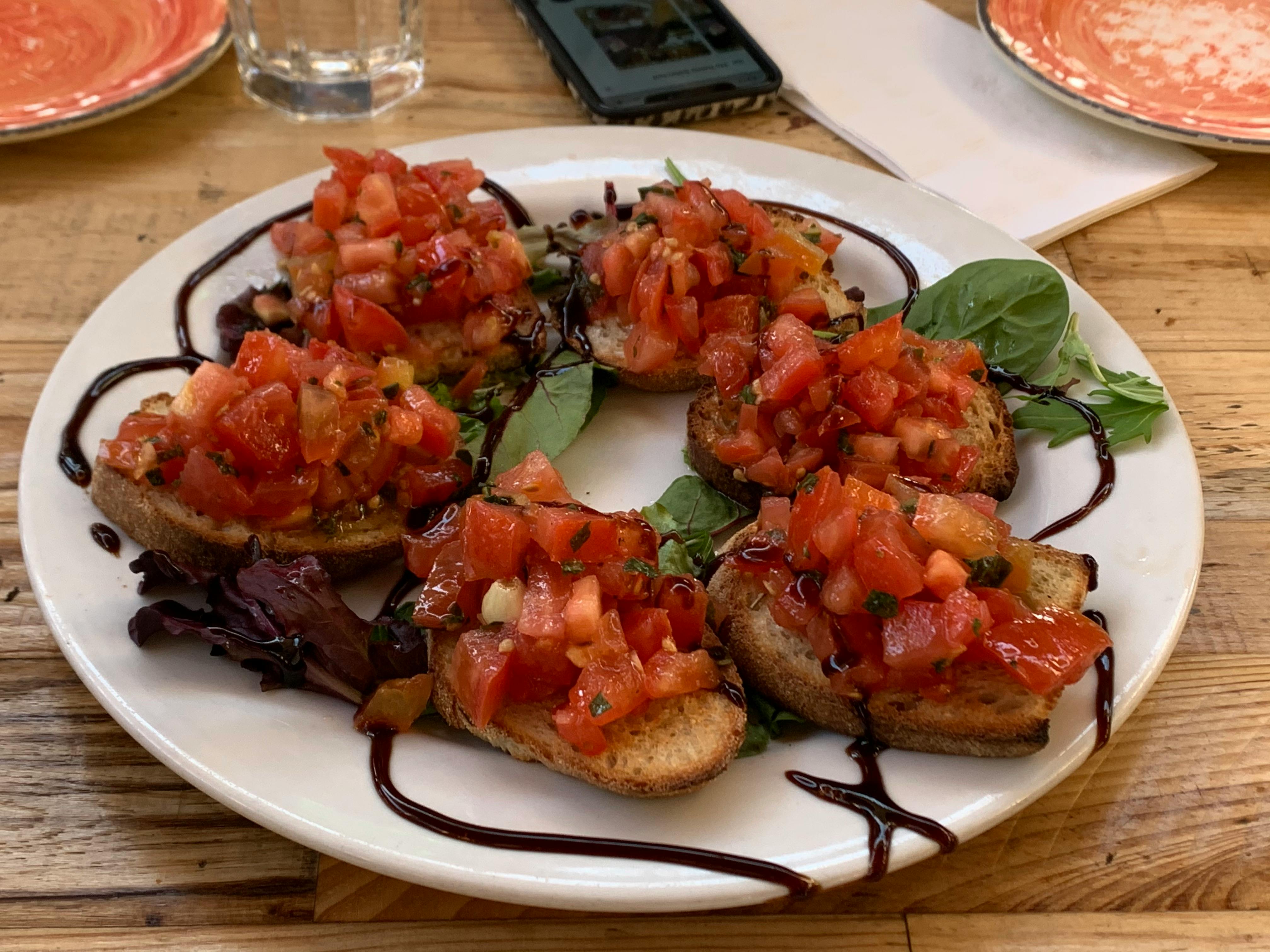 A plate of bruschetta topped with diced tomatoes and garnished with greens, drizzled with balsamic glaze. A phone and glass are in the background.