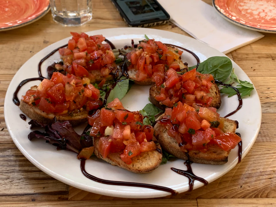 A plate of bruschetta topped with diced tomatoes and garnished with greens, drizzled with balsamic glaze. A phone and glass are in the background.