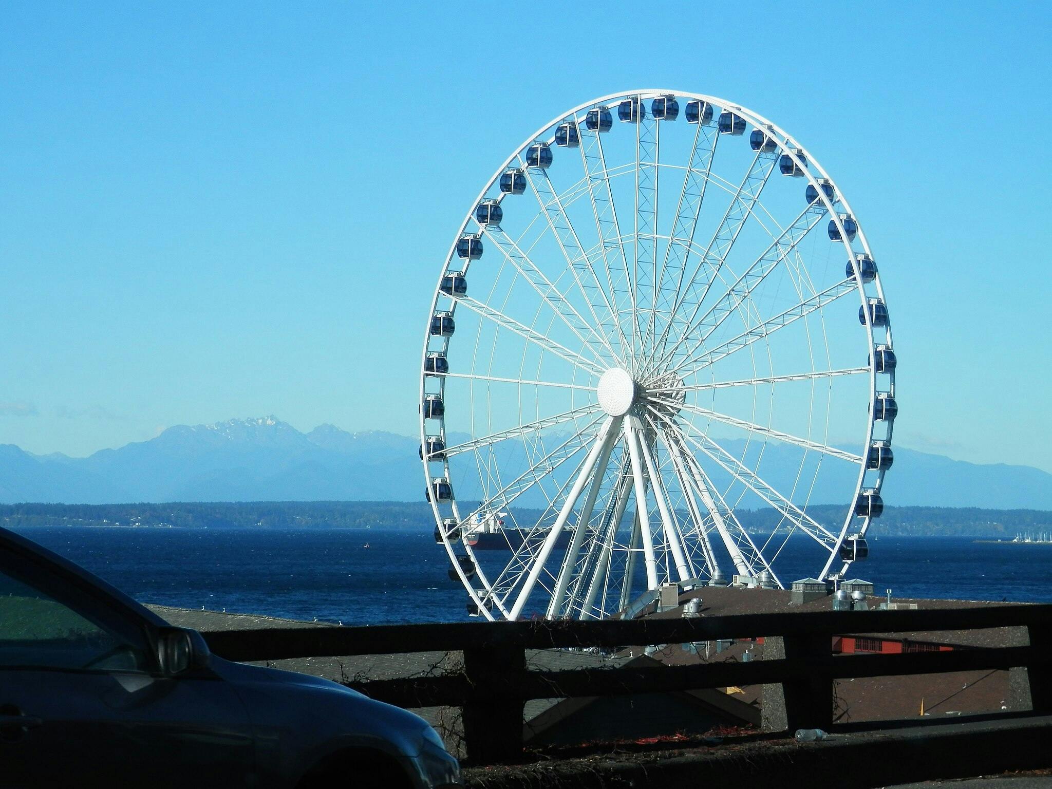 Une grande roue au bord de la mer avec des montagnes en arrière-plan. Une voiture est partiellement visible au premier plan.