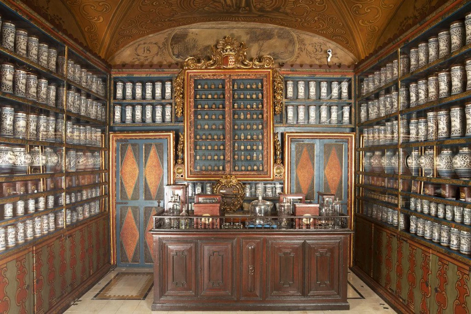 Vintage apothecary with wooden cabinets, jars lining the shelves, and a counter displaying various bottles and containers.