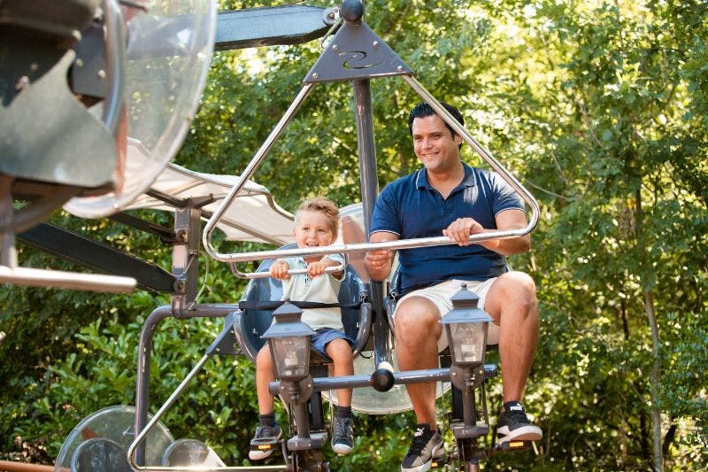 A man and child smile while riding an amusement park bicycle carousel. Trees are visible in the background.