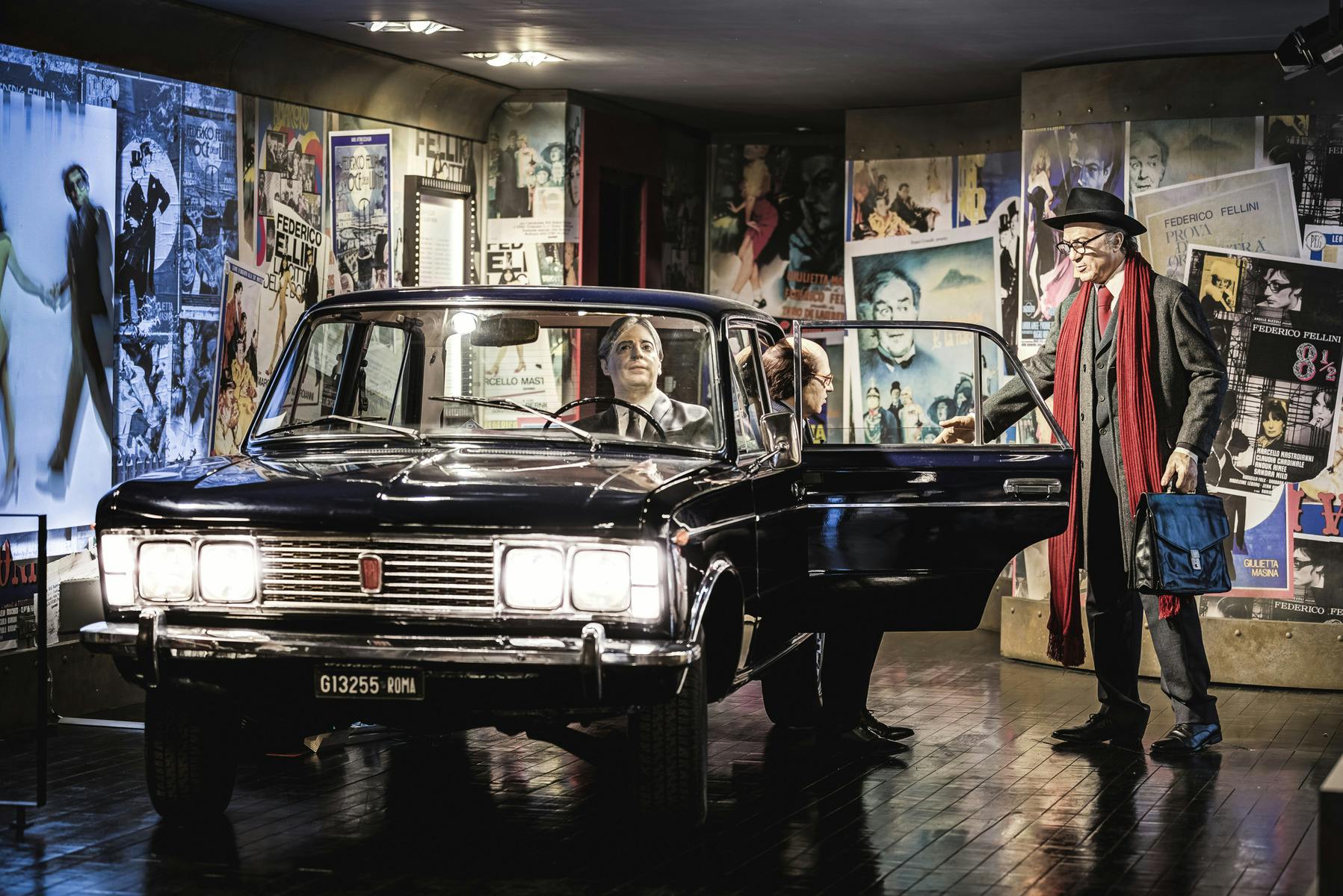 Un uomo con cappello e sciarpa è in piedi accanto a un'auto d'epoca nera, con un altro uomo seduto all'interno. Le pareti sullo sfondo sono ricoperte di manifesti.