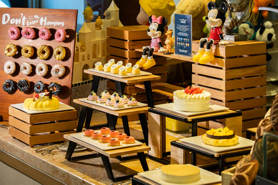 A dessert display with cakes, pastries, and Disney-themed decorations on tiered wooden stands in a bright setting.