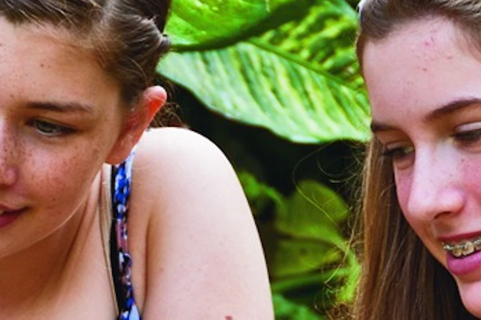 Two young people kneeling on grass feeding a small kangaroo in a lush outdoor setting with green foliage.