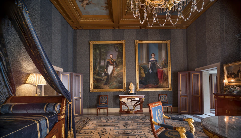 Ornate room with large framed portraits, intricate ceiling, chandelier, detailed rug, wooden furniture, and a marble console table.