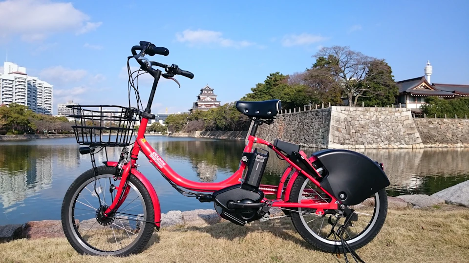 A red electric bicycle parked near a calm river with a traditional Japanese castle and trees in the background.