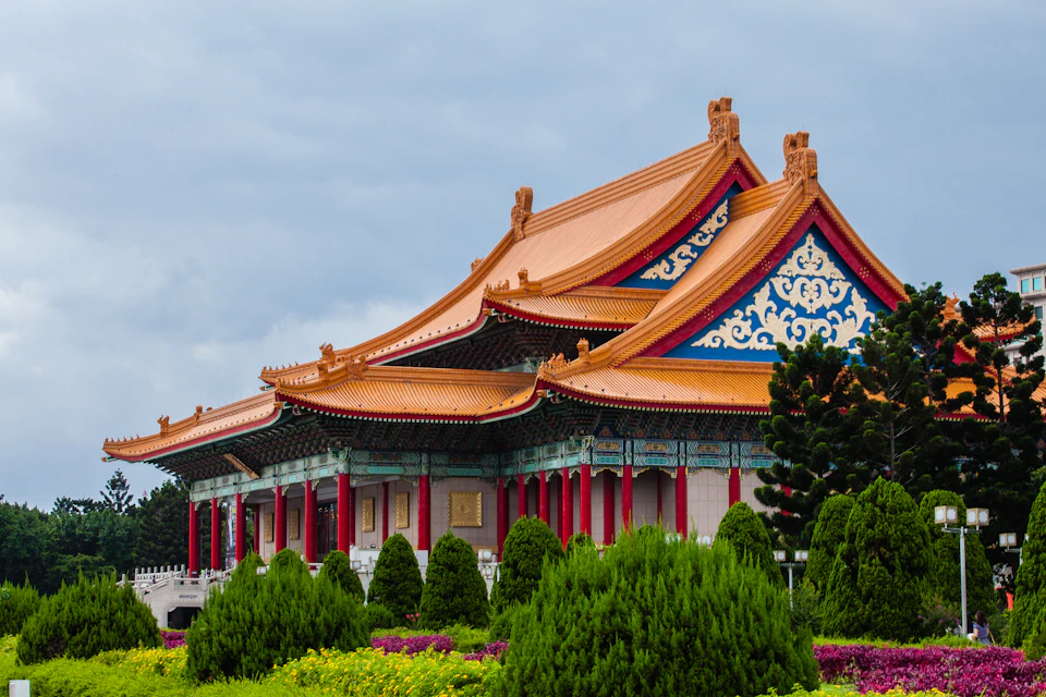 Traditional Chinese building with red pillars, ornate blue detailing, and orange curved roofs, surrounded by green shrubs and flowering plants.
