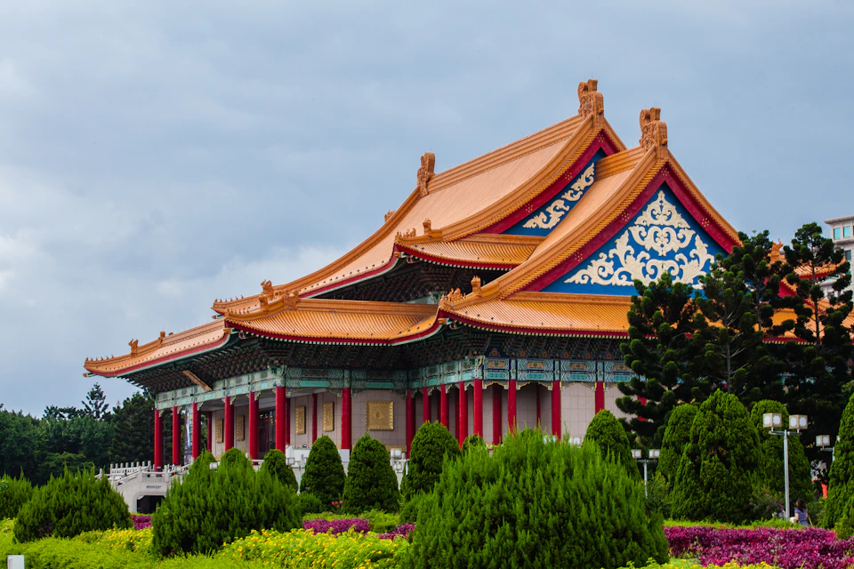 Traditional Chinese building with red pillars, ornate blue detailing, and orange curved roofs, surrounded by green shrubs and flowering plants.