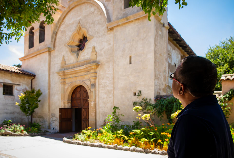 A person looks at a historic stone building with a large wooden door, arched window, and surrounding greenery under a clear sky.