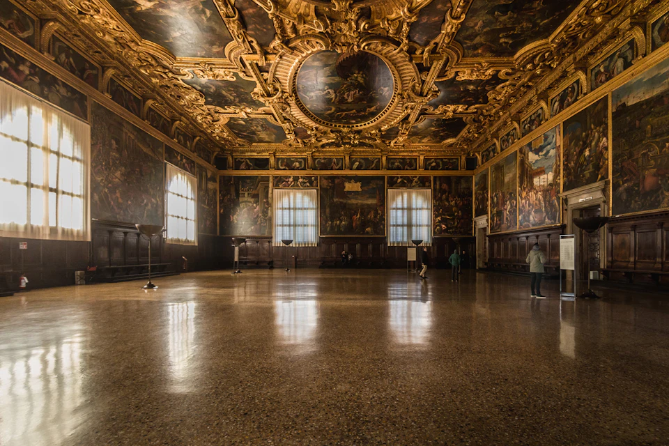 Grand hall with ornate gold and painted ceiling, large windows, and people observing wall art. Floor is polished and reflective.