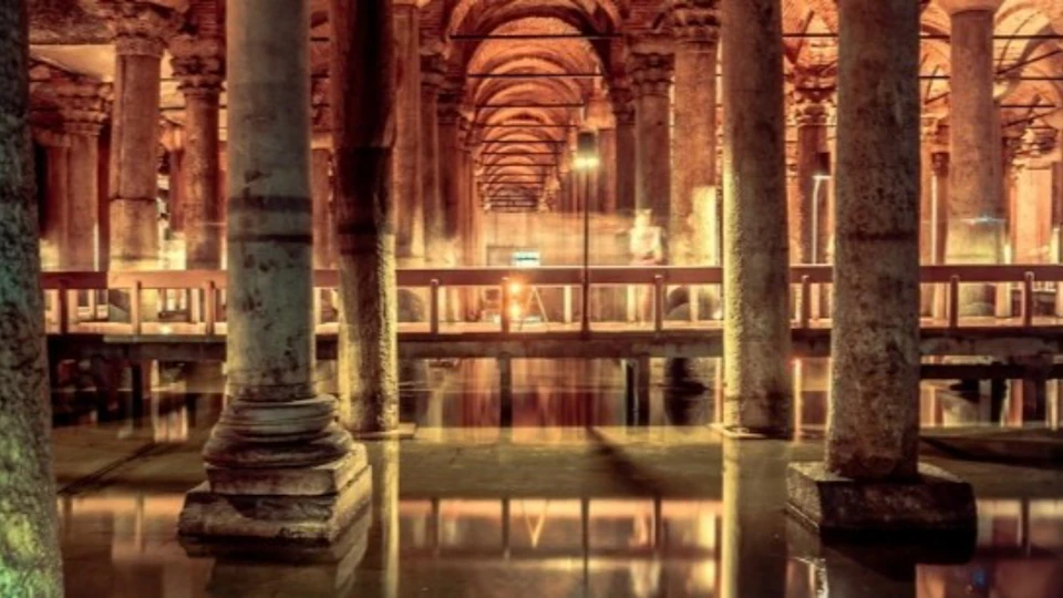 A dimly lit underground structure with stone columns and arches reflected in water, illuminated by warm lighting.