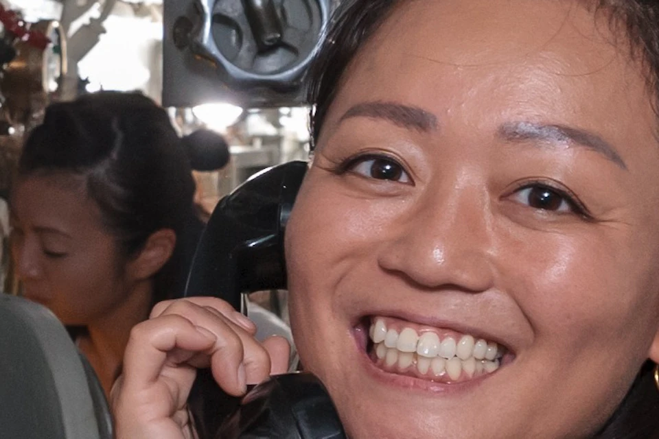 A person smiling while holding a phone receiver, standing in a submarine's interior with visible equipment and pipes.