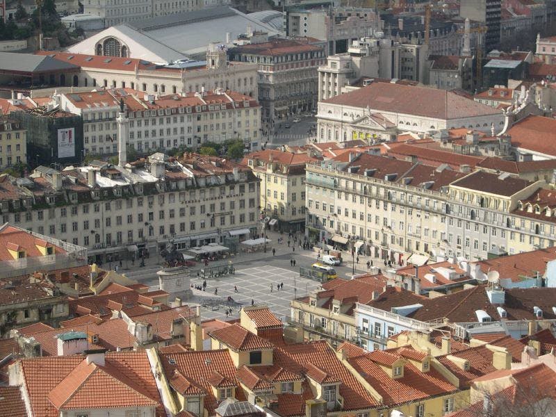 Flygfoto över ett torg omgivet av historiska byggnader med röda tak; några personer och fordon är synliga på torget.