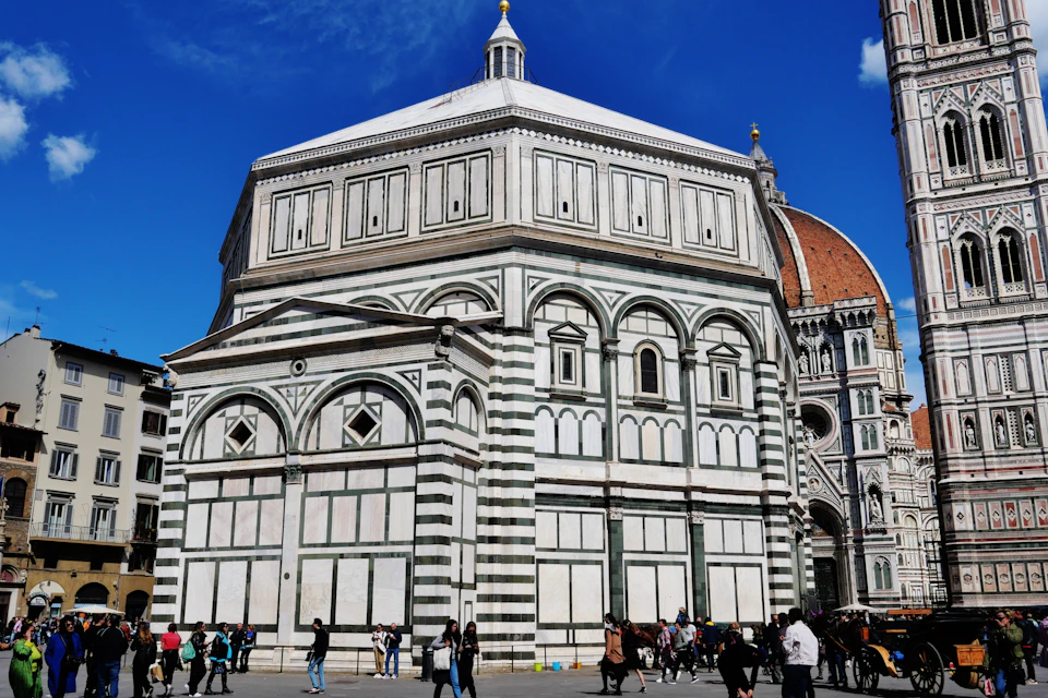 A white and green octagonal building with ornate patterns and a dome in the background, with people walking around it.
