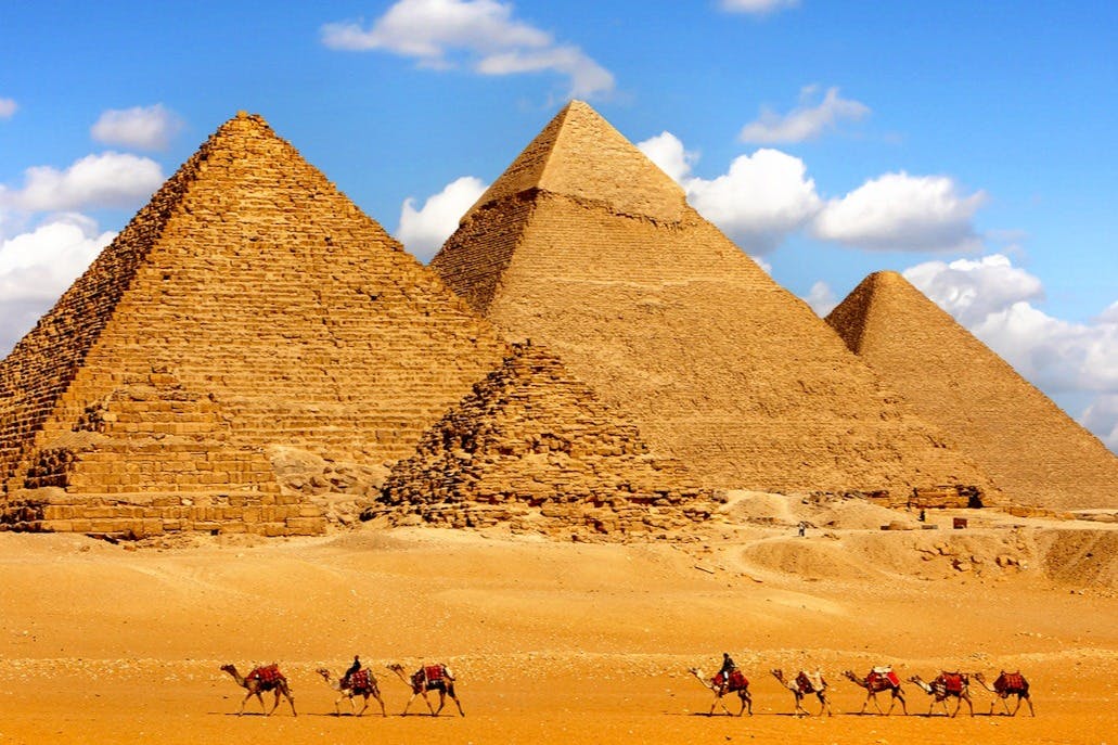 Οι Πυραμίδες της Γκίζας κάτω από έναν καταγάλανο ουρανό, με καμήλες και αναβάτες που ταξιδεύουν σε όλη την έρημο σε πρώτο πλάνο.