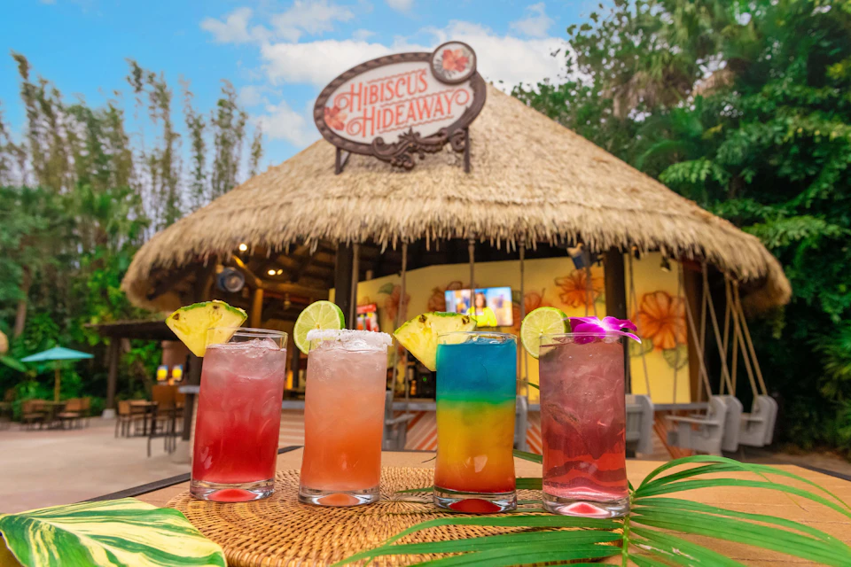 Four colorful cocktails with garnishes in front of a tiki bar named Hibiscus Hideaway, under a clear sky.