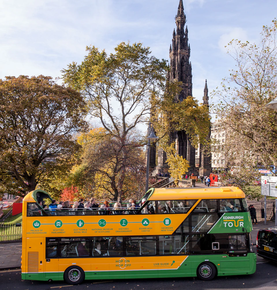 A yellow and green double-decker tour bus passes by trees and a historic gothic monument in an urban park setting.
